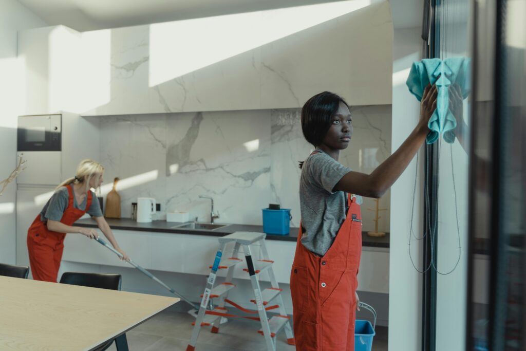 Two women cleaning a modern kitchen, wiping glass surfaces and mopping the floor.